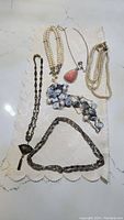 Group photo of five different necklaces laid on a white lace cloth, showing variety including pearl strands, a coral teardrop pendant, a chunky blue and white stone necklace, and two metallic chain necklaces.