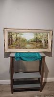 Full view of framed oil painting of spring landscape on a stand against wall.