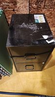 Top view of the small black metal filing cabinet showing two drawers and stickers residue on top.
