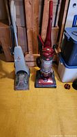 Overview photo showing both the Bissell carpet cleaner and the Hoover upright vacuum side by side on a yellow floor against wooden panel background.