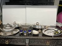 Wide view of the lot showing all silver plated serving dishes arranged on a carved dark wooden surface. Includes soup tureen, covered dishes, small trays and egg cups.