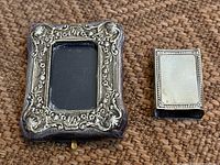 Photo showing antique sterling silver picture frame with ornate floral and scrollwork border, beside a sterling silver matchbox cover with engraved border.
