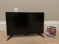 Front view of Insignia 22 inch LCD TV with black frame and two black angled legs on a carpeted floor against beige wall. To the right, boxed George Weasley Funko Pop! figurine visible.