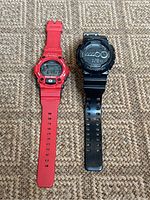 Pair of Casio G-Shock watches, red G-7900A and black GD-100, side by side on carpeted surface.