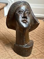 Full front view of bronze head sculpture on carpeted floor against white wall.