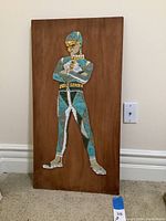 Full view of the wood panel with the stone mosaic figure on a beige carpet against a white wall.