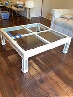 White wooden frame square coffee table with four clear glass panels on top, placed on a wooden floor next to a white tufted sofa and lamp.