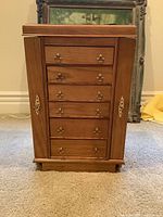 Front view of wood jewelry armoire cabinet showing six drawers and two side doors with brass handles and accents.