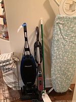 Wide shot showing Oreck vacuum, Shark steam mop, green handle mop, metal corner shelf, ironing board with floral cover, and mop heads on the shelf.