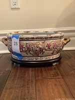 Front and side view of the oval porcelain pot featuring floral patterns with gold handles and resting on a wooden base.