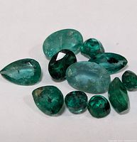 Ten loose emerald gemstones arranged on a white surface showing various shapes and green tones.