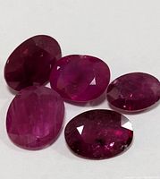 Five oval faceted genuine ruby gemstones displayed on a white background showing rich varying red shades and clarity variations.