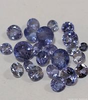 Close-up photo showing multiple round faceted tanzanite gemstones with light to medium violet-blue color on a white reflective surface.