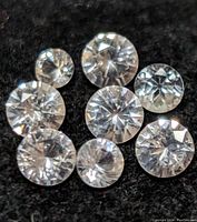 Photo showing nine loose round brilliant cut white sapphire gemstones arranged on a dark fabric background.