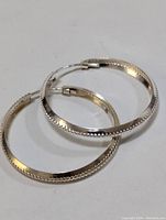 Pair of silver hoop earrings arranged overlapping each other on a white background, showing the texture on the edges and clasp detail.
