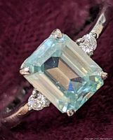 Close-up image of emerald cut light greenish-blue moissanite stone set in a sterling silver band with two round small clear accent stones on either side.
