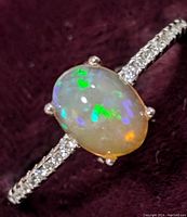 Close-up image of sterling silver ring featuring an oval opal with green, blue, and orange flashes, and a band studded with small round cubic zirconia stones.