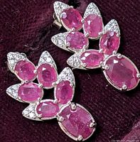 Close-up photo of pair of earrings showing multiple oval and pear-shaped pink rubies surrounded by small white cubic zirconia stones on a dark background.