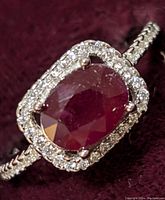 Clear close-up photo showing oval ruby center surrounded by sparkling cubic zirconia stones, detailed band visible.