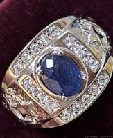 Close-up photo showing the oval faceted blue sapphire center with surrounding clear cubic zirconia stones and star decorative accents on the sterling silver band.