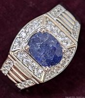 Close-up front view of the sterling silver ring featuring a blue sapphire center stone surrounded by clear cubic zirconia stones and ribbed band design with additional stones.