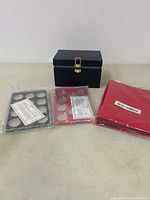 Two coin holders (black and red), black RFID key box with gold latch, and a packaged coin collection book on a table.