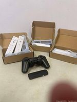 Three white Nintendo Wii Remotes with wrist straps and silicone sleeves in open boxes, three white Wii Nunchuks, one black PS3 controller, and one PS3 Bluetooth dongle visible on white background.