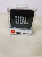 Photo showing JBL GO 2 Bluetooth speaker in its sealed transparent plastic packaging with black speaker and JBL logo visible.