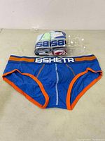 Blue men's briefs with orange trim and BSHETR branding on the waistband laid flat.
