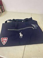 Navy blue Ralph Lauren Polo duffle bag lying flat on a light colored surface showing front zipper pocket, white embroidered Polo player logo, red Polo shield patch, and embroidered 'P' with wings.