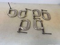 Six stainless steel C-clamps arranged in two rows on a flat surface showing overall condition and size variation.