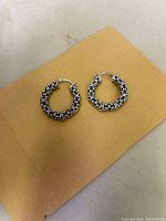 Pair of sterling silver Byzantine hoop earrings displayed on beige paper background showing detailed pattern and latch closure.