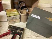 Box containing multiple rolls of tape including carpet tape in original box, masking tape, duct tape, and kraft paper tape with a metal staple gun beside it on a wooden surface.