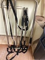 Two metallic bed rails with black padded grips and black straps, leaning against a wall and door. The floor is wooden and part of a rolled carpet is visible.