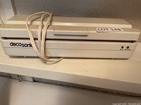 White Decosonic heat sealer unit with power cord coiled on top. Shows front of unit and brand label.