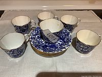 Overview of six blue floral bone china teacups and six matching saucers arranged on white cloth.
