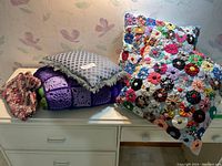 Set of colorful handmade cushions, purple crocheted afghan blanket, and floral fabric pouch displayed on a white piece of furniture.