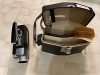 Image showing the movie camera next to an open black carrying case containing manuals and papers.