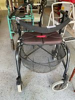 Front view showing teal and black walker frame with brown padded seat, black curved support bar, and front wheels.