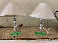 Pair of small table lamps with tassel-shaped clear glass and metal bases, fabric lampshades, placed on a carpeted floor.