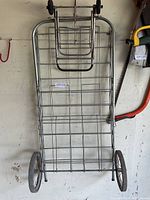 Metal shopping cart with wire mesh frame, large wheels, and handlebar leaning against a wall