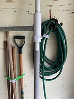 Photo showing long white Super Pole with attached Super Bar, green garden hose coiled next to wall, three wooden-handled garden tools leaning against wall.