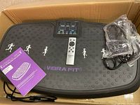 Vibra Fit vibration platform showing textured black surface with running figures, control panel, remote control, and user manual in original box.