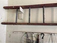 Side view of wooden ladder with red rails and metal rungs hung on garage wall with other tools visible below