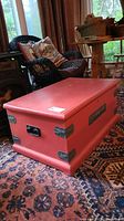 Solid wood travel trunk shown from side angle in a room with wicker chair and carpet visible. Painted red with black metal handles and green corner brackets.
