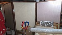 Photo of multiple pegboard display dividers framed in wood stacked upright in a basement storage area. Shows condition and finish on boards and frames, with some objects nearby for scale.