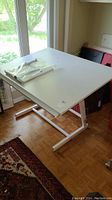 White drafting table with adjustable surface angle and metal frame base, shown near window in room with parquet floor.