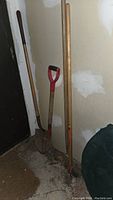Photo showing wood-handled shovels and a rusty pitch fork against a wall.
