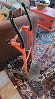 Full view of orange frame hand cart dolly with black handles and wheels placed indoors on rug.