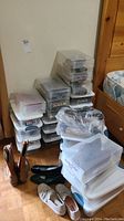 Wide view of many clear plastic shoe storage boxes stacked with some shoes on the floor next to them.